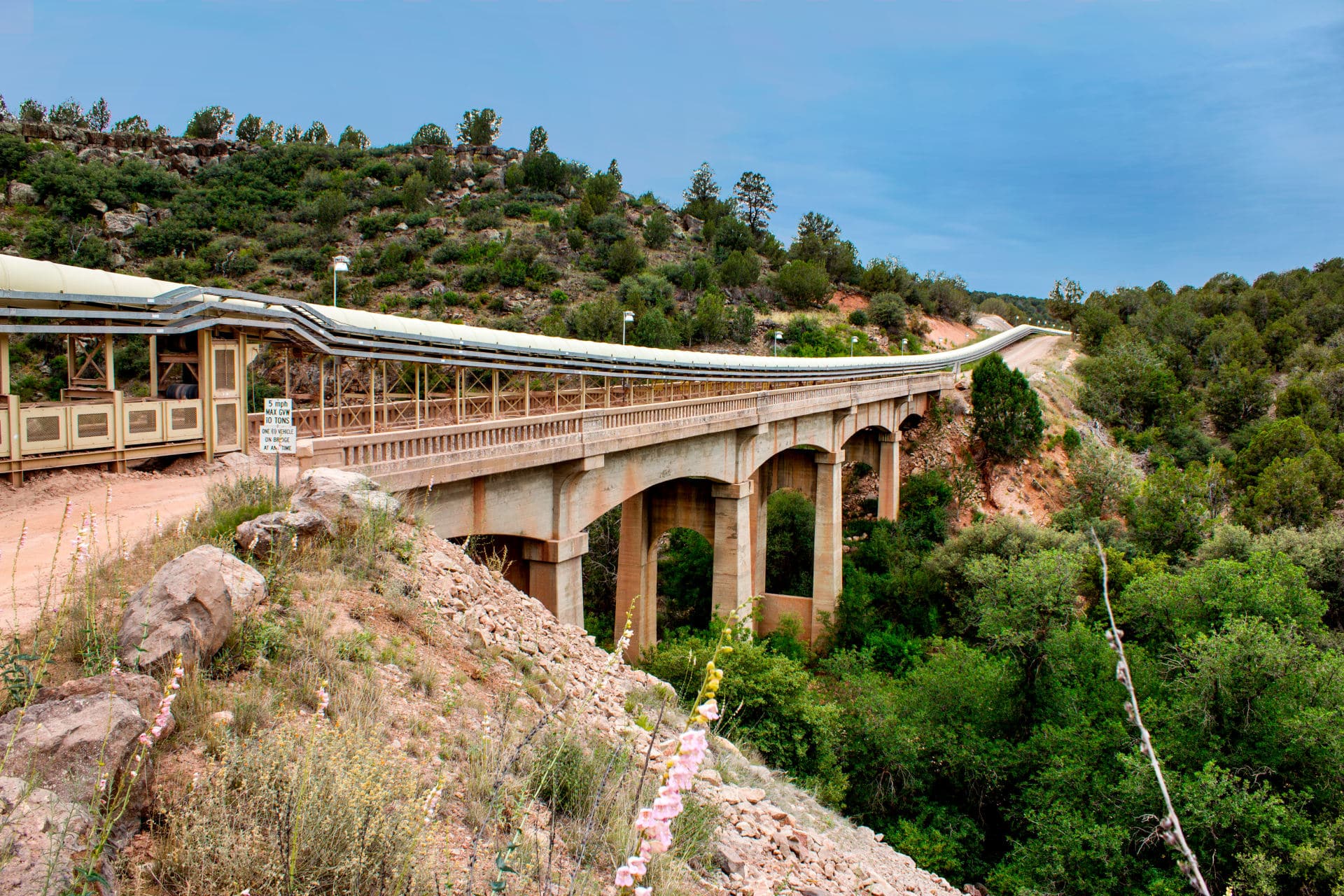A covered conveyor bridge at the Drake Cement facility in Paulden, Arizona, spanning a canyon with surrounding vegetation and hills.