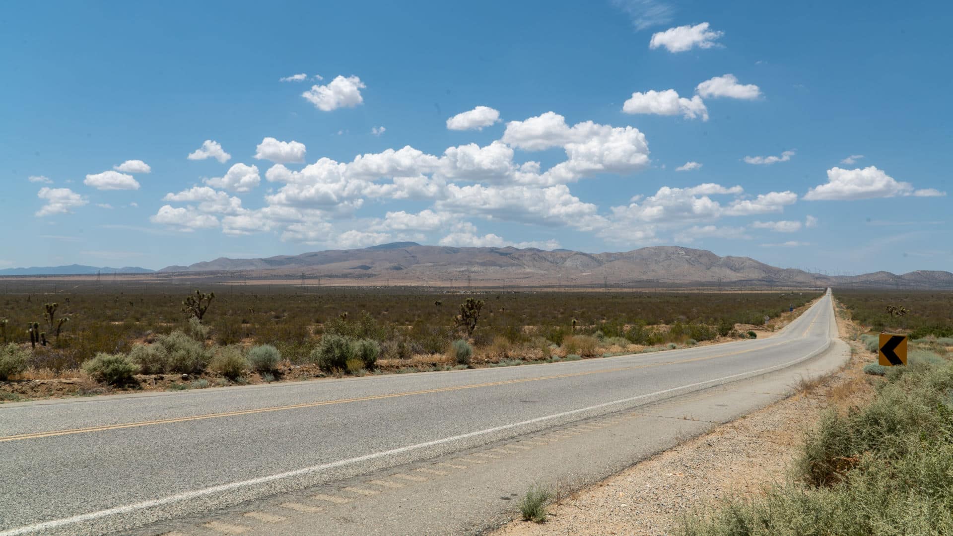 A long desert highway leading toward the mountains near Tehachapi, California, under a bright sky with scattered clouds.