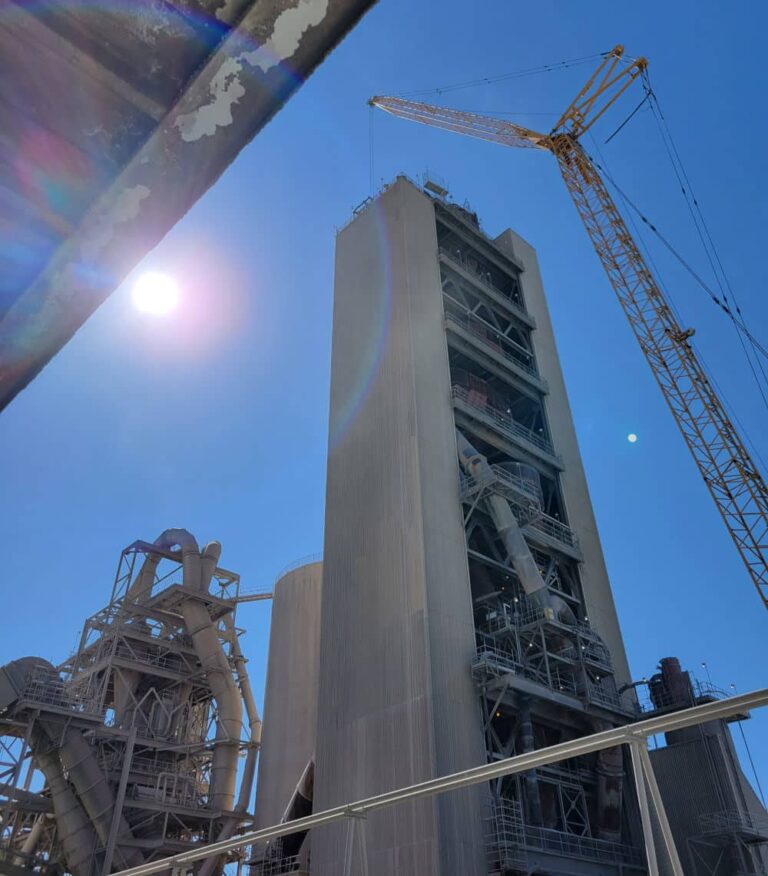 A tall industrial tower and supporting structures at the Tehachapi Cement plant with a crane overhead under a bright blue sky.