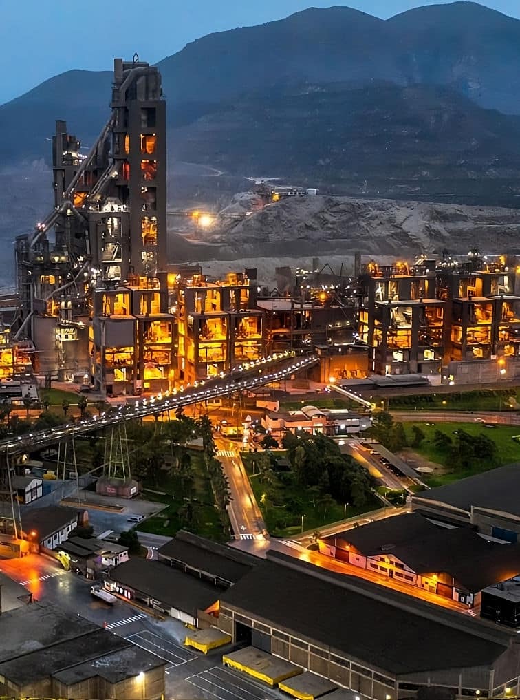 A brightly illuminated cement manufacturing plant at night with conveyors, industrial towers, and surrounding mountain landscape.