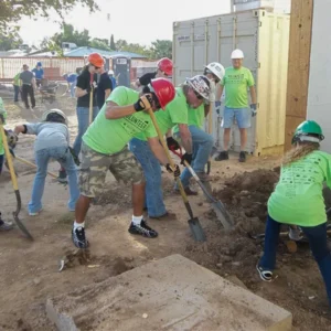 A group of volunteers wearing safety gear and green shirts working together on a community improvement project with shovels and tools.