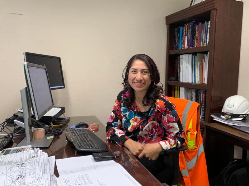 A UNACEM North America employee sitting at a desk in a corporate office with computer monitors, documents, and safety gear in the background.